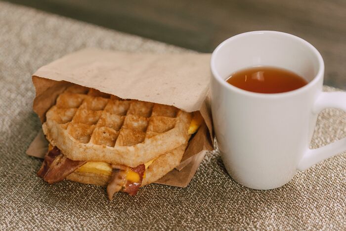 Waffle sandwich with bacon and egg next to a white mug filled with tea on a textured surface, bite my nails concept.