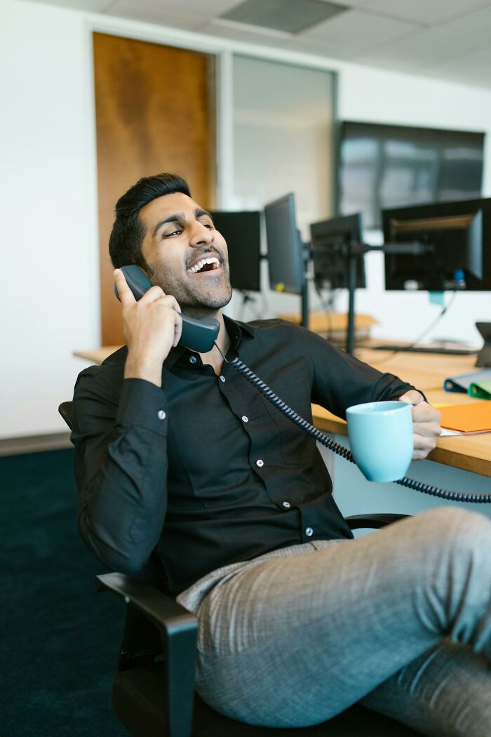 Male coworker laughing on phone at office desk holding coffee mug in a casual workplace setting.