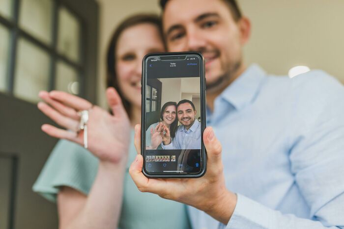 A happy couple holding house keys and showing a selfie on phone, illustrating hidden costs of buying a home.