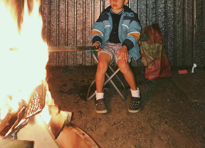 Child sitting by a campfire at night, wearing a jacket and shorts, capturing an intense hospital experiences vibe.