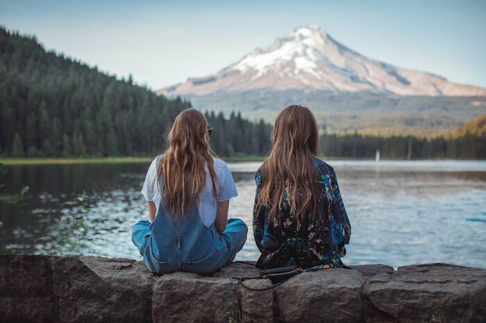 Two people sitting on a stone wall by a lake, surrounded by nature, reflecting on personal mysteries and human nature.