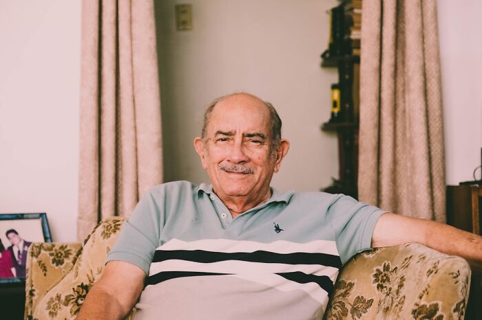 Elderly man sitting on a patterned couch, smiling comfortably, sharing his best I was there when it was written moment.