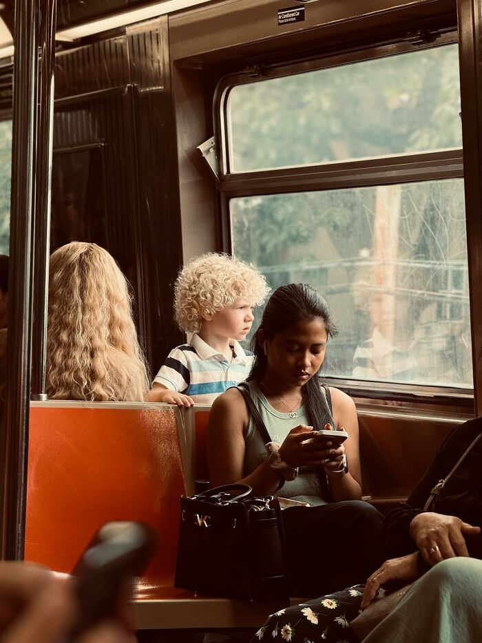 Child with curly hair looking outside the window on a bus while a woman nearby focuses on her phone, illustrating complex parent-child dynamics.