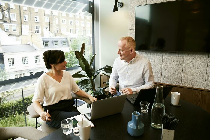 Two colleagues discussing company knowledge loss in a modern office with laptops and a large screen TV nearby.