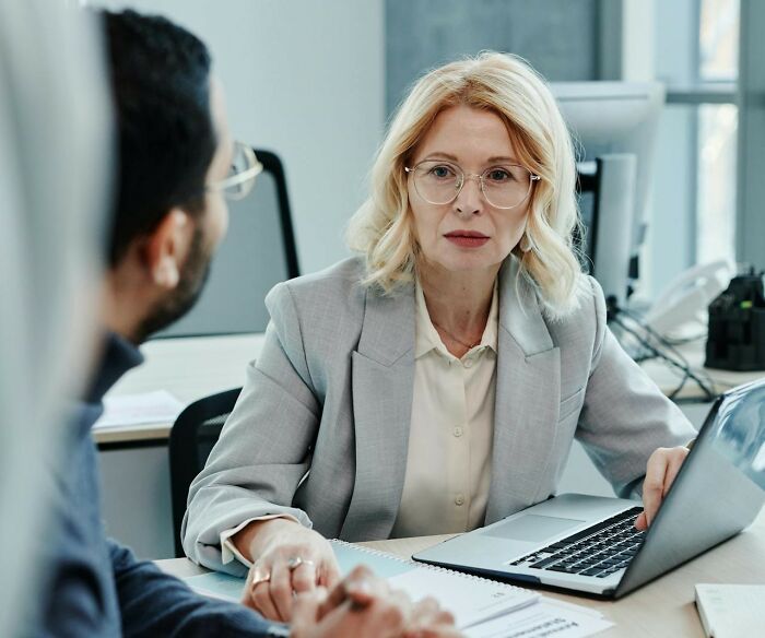 Woman over 42 in a gray blazer having a serious conversation with a colleague in a modern office setting.