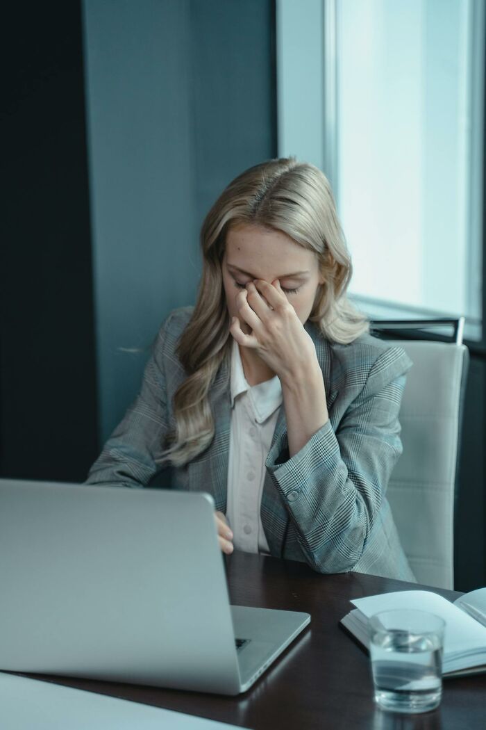 Stressed woman in a gray blazer at her laptop experiencing frustration from male coworkers in a professional office setting.