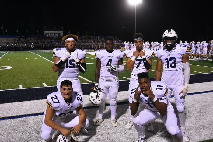 Six high school football players in white uniforms pose on the field at night, showing pride in their glory days.