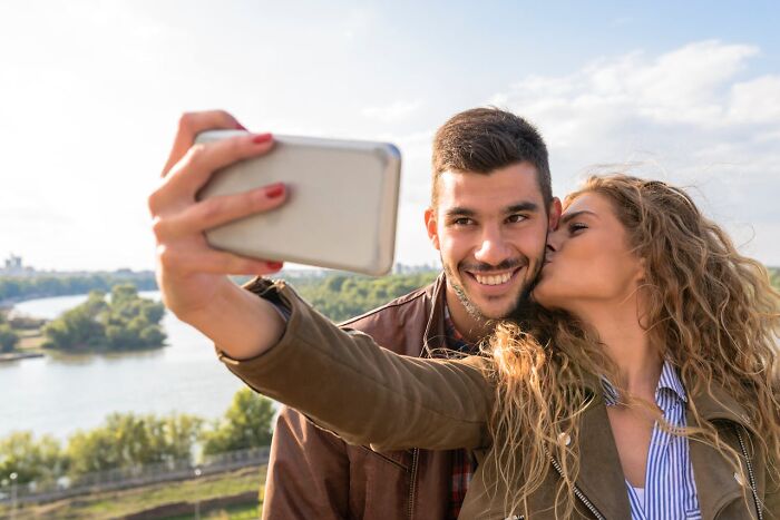 A woman kissing her partner on the cheek while taking a selfie outdoors illustrating wildest things exes get away with.