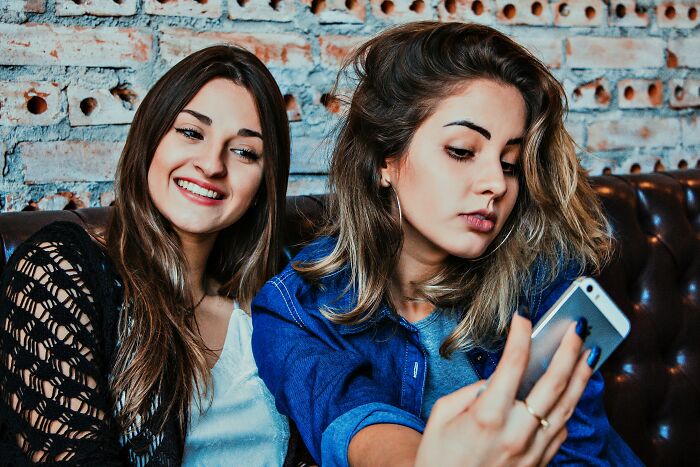 Two young women sitting on a couch, one smiling and the other showing subtle signs of low self-esteem while looking at her phone.