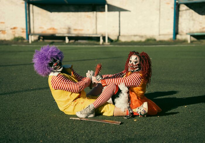 Two scary clowns in colorful costumes and creepy makeup playfully wrestling on a grassy field in daylight.