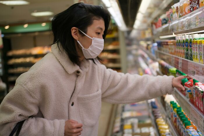 Young woman wearing a mask while shopping, reaching for a drink among refrigerated shelves, reflecting a completely legal total psychopath theme.