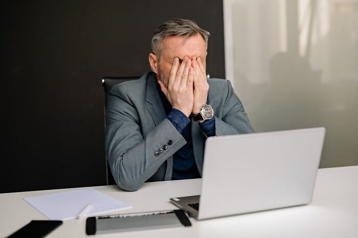 Man in a gray blazer covering his face while sitting at a desk with a laptop, reflecting on I was there when it was written moments.