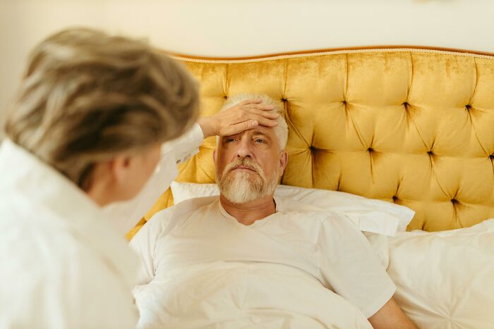 Older man lying in bed looking unwell as a woman checks his forehead, depicting coworker diabolical things reported to HR.