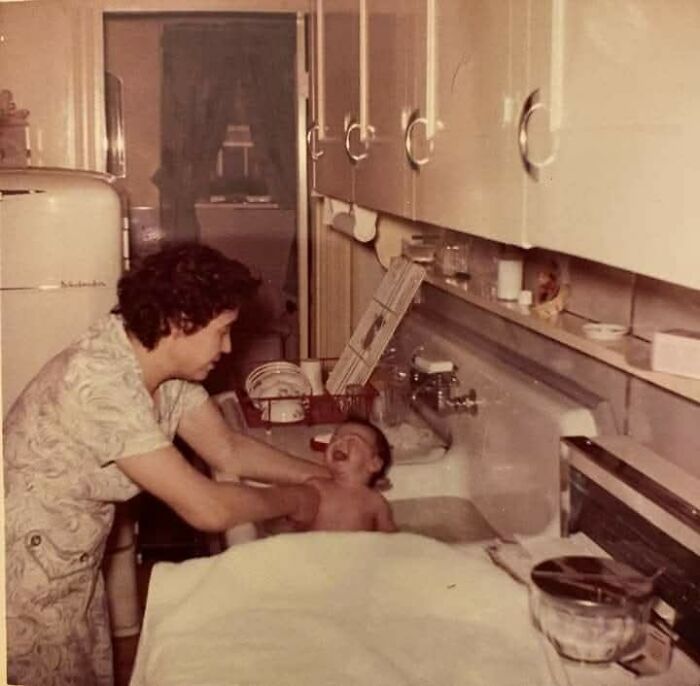 Vintage photo of a mother bathing her baby in a kitchen sink, showing moments from the past.