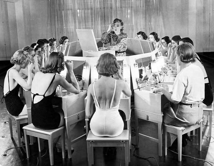 Women seated around a circular makeup station applying cosmetics in a timeless photo capturing what the past looked like.