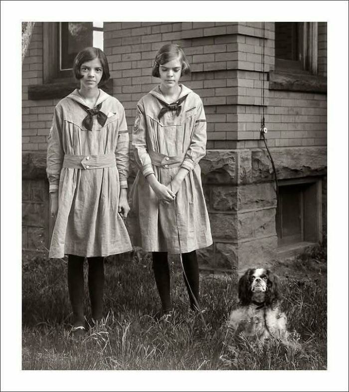 Two girls in vintage dresses standing with a dog outside a brick building in timeless photos showing the past.