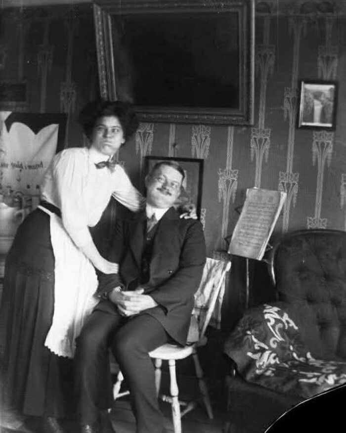 Black and white photo showing a man sitting and a woman standing, capturing a timeless moment from the past.