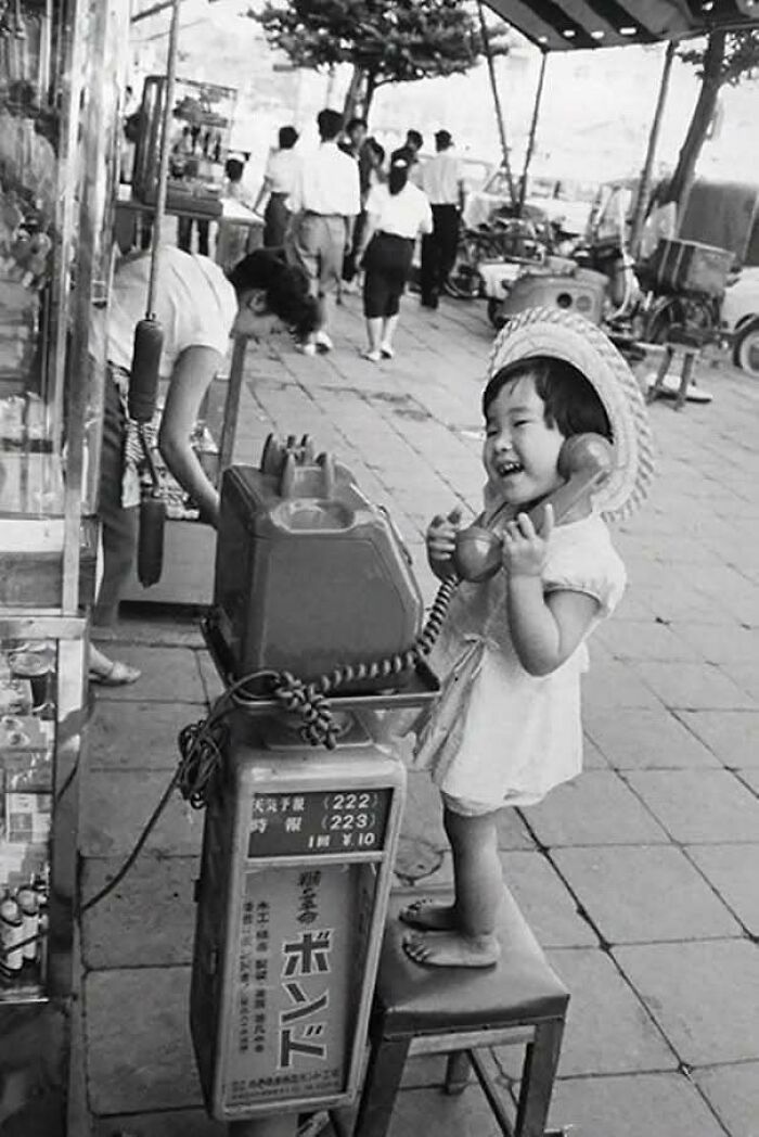 Niña sonriente usando un teléfono público vintage en la calle, mostrando un lado distinto de la historia antigua.