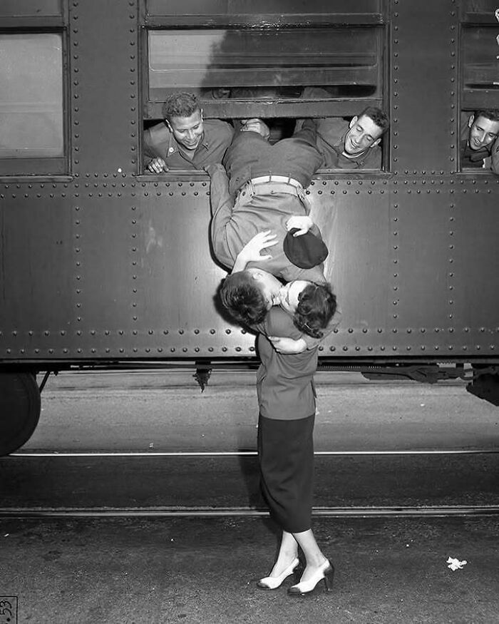 Soldier kissing a woman while hanging out of a train window, with friends watching, a timeless photo showing the past.