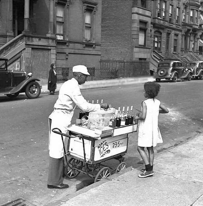 Black and white photo of a vendor selling ice treats from a street cart to a young girl, showing the past life scene.