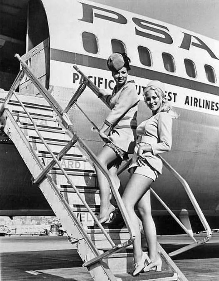 Two vintage airline stewardesses smiling on stairs boarding Pacific Southwest Airlines plane in a timeless photo from the past.