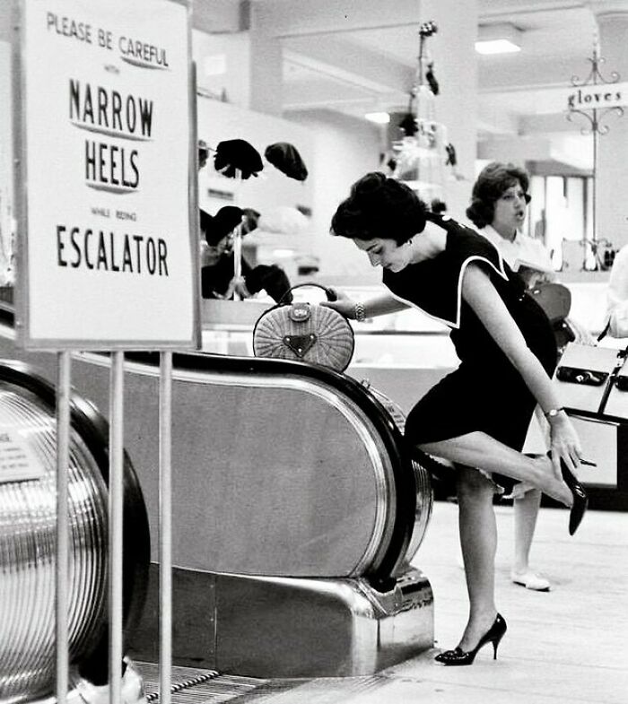 Mujer con tacones altos ajustándose el zapato junto a una escalera mecánica, foto histórica mostrando un lado distinto de la historia.