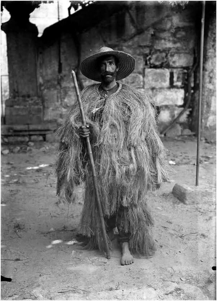 Man wearing traditional straw costume and wide-brimmed hat holding a staff in a timeless photo showing the past.