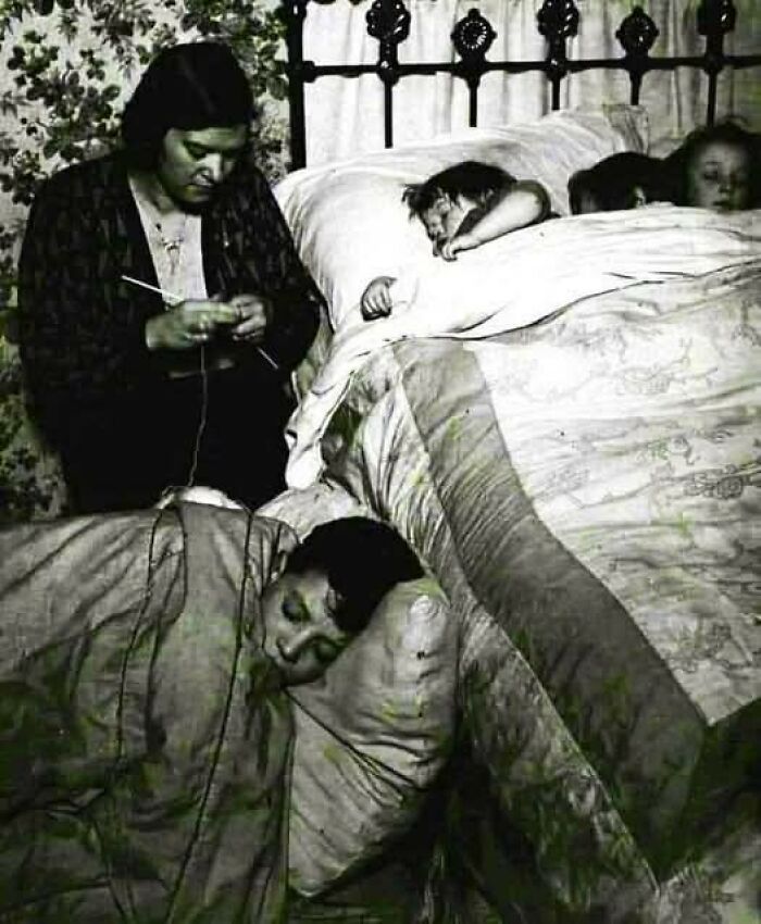 Woman knitting beside children sleeping on bed and floor in a cozy room, vintage black and white timeless photo from the past.