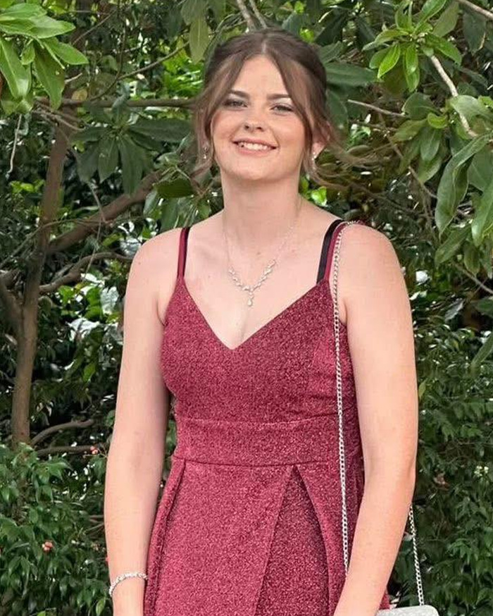 Teen mourner in a red dress standing outdoors surrounded by greenery, related to hearse and funeral events.