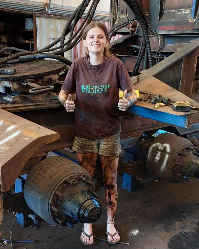 Teen mourner showing thumbs up, covered in dirt, standing in a workshop near heavy machinery parts and equipment.