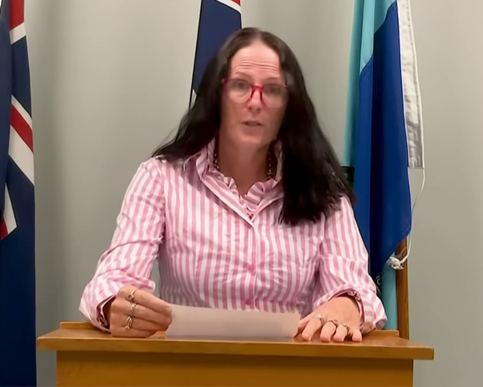 Woman with long dark hair and red glasses speaking at a podium with flags behind her in a formal setting