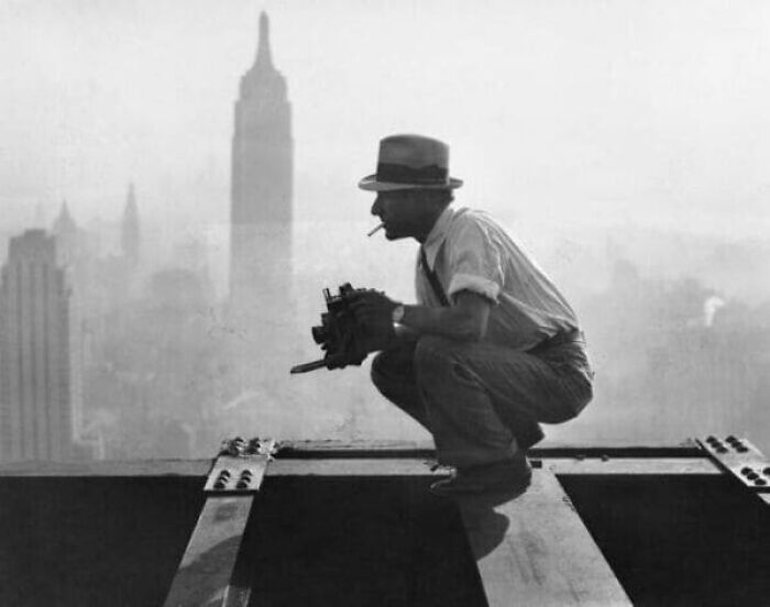 Man with camera and cigarette crouches on construction beam with city skyline in background, photos alternative angles unseen.