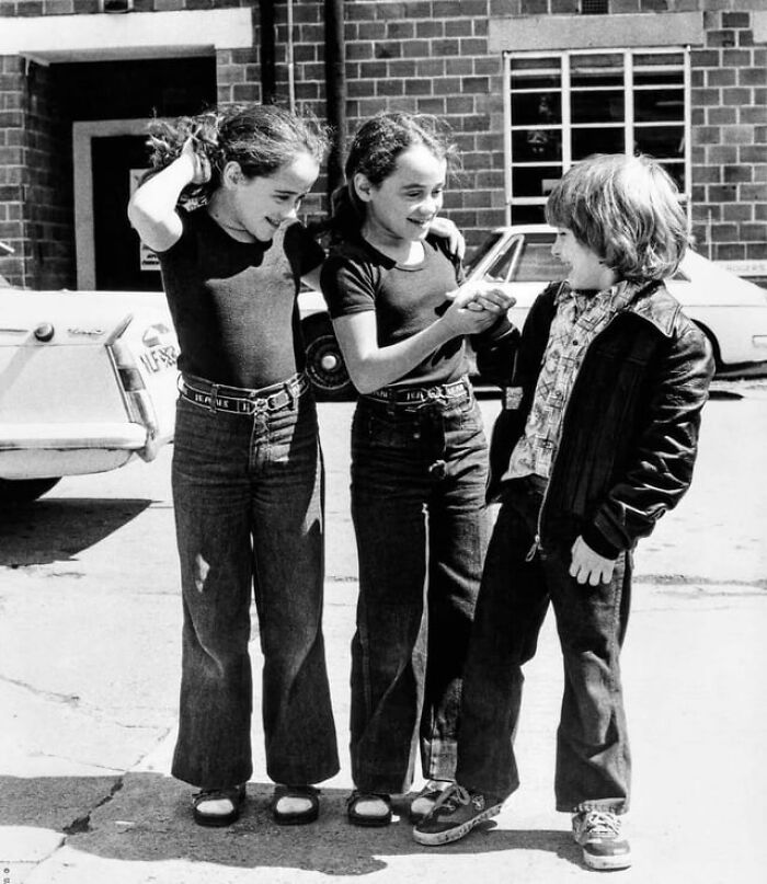 Two girls and a boy laughing and interacting outdoors, captured from alternative angles in an unseen moment.