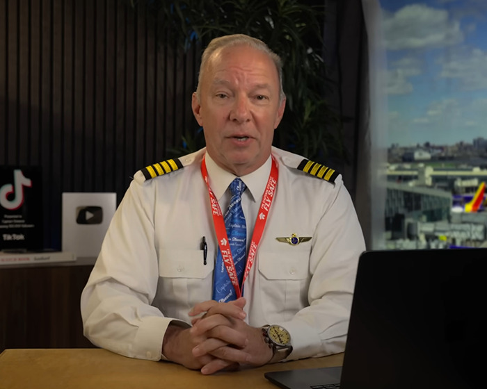 Pilot in uniform with Air India badge seated at desk, discussing new enhanced clip before plane crash investigation.