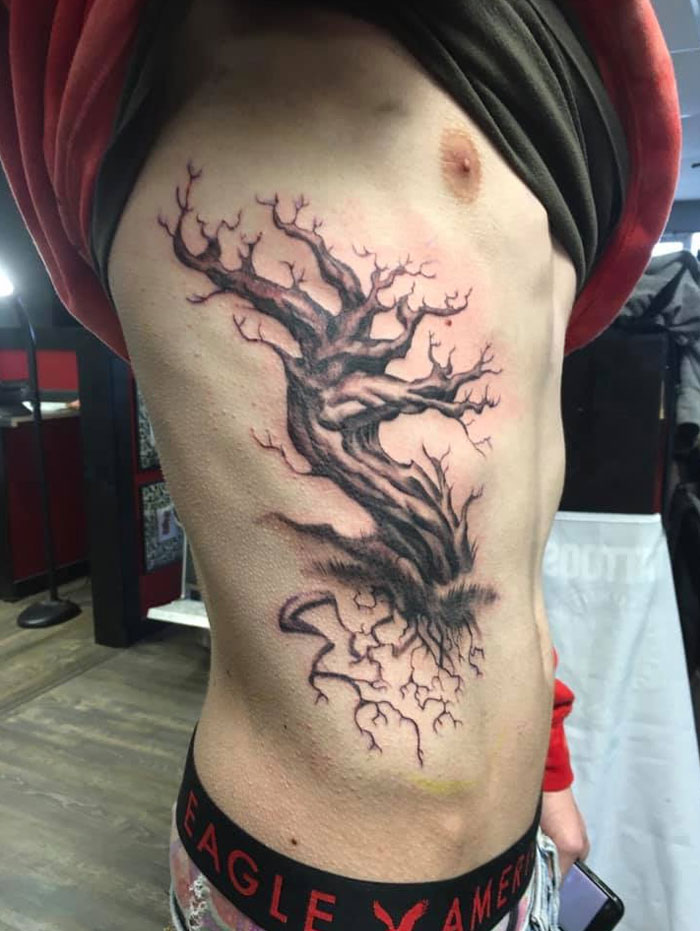 Black poison tree tattoo design with twisted branches and roots on person's side torso in a tattoo studio setting