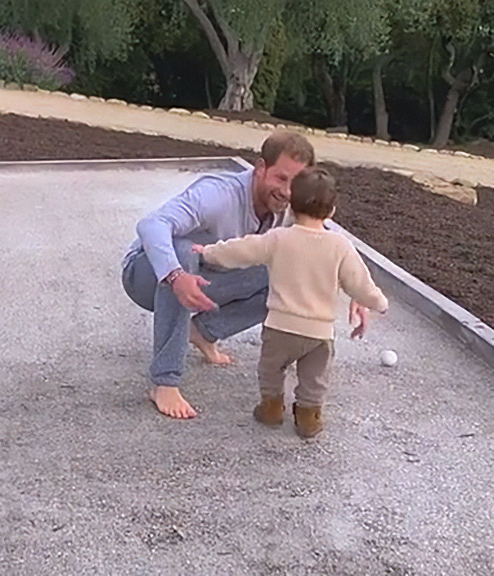 Man crouching barefoot on gravel with toddler, related to Meghan Markle Father's Day post and paid child actors controversy. Man crouching barefoot on gravel with toddler, related to Meghan Markle Father's Day post and paid child actors controversy.