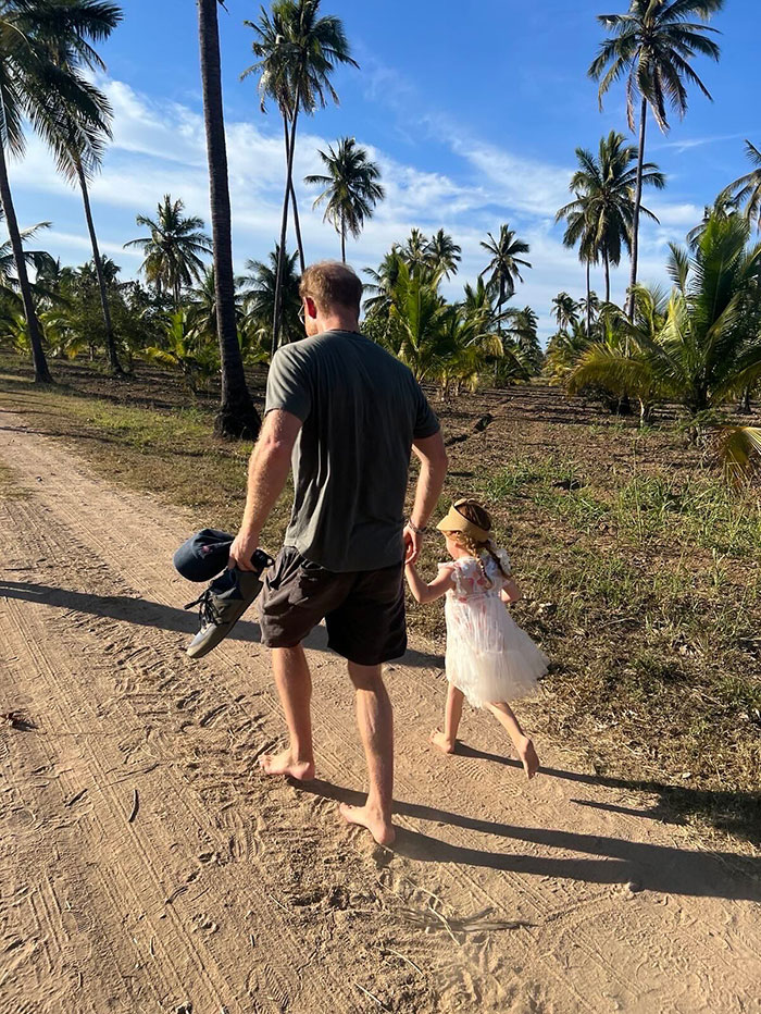Man and little girl walking barefoot on a dirt path lined with palm trees, sunlight casting long shadows at sunset.