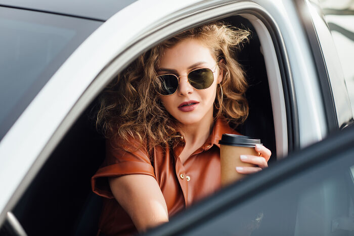 Young woman with sunglasses in a car holding a coffee cup, reflecting rich kid meltdowns at private schools.