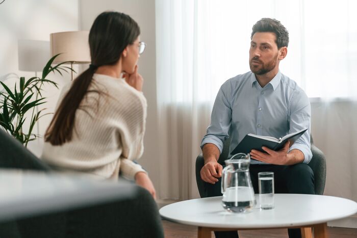 Therapist in a blue shirt holds a notebook while listening intently to a woman during a confidential therapy session.