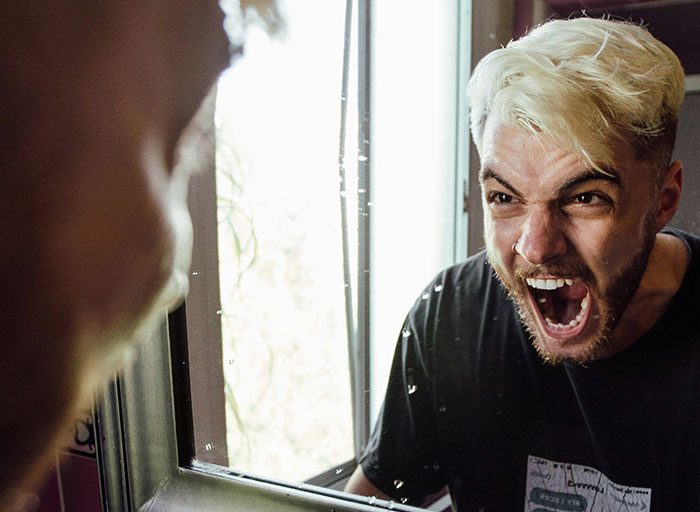 Angry young man with blond hair screaming at his reflection in a mirror, capturing intense emotion and fear in psychology context.
