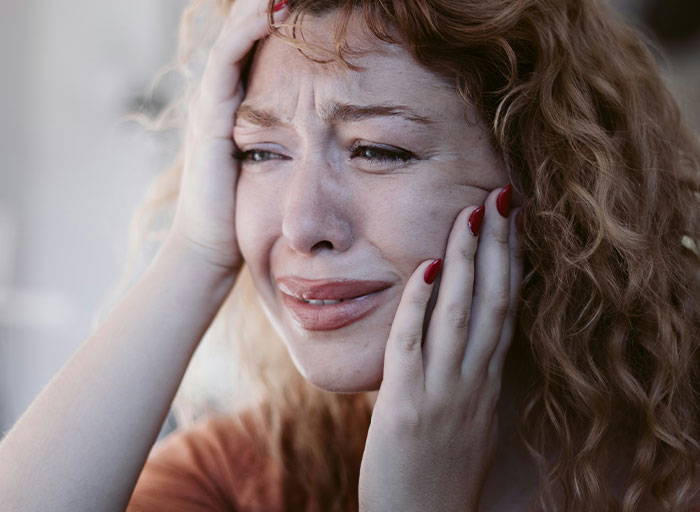 Woman with red hair and nails holding her head, crying and showing signs of emotional distress in a close-up portrait.