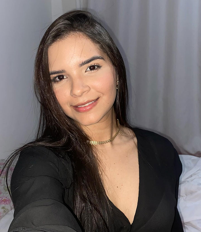 Young beauty queen smiling softly, wearing a black top and gold necklace in a softly lit indoor setting.