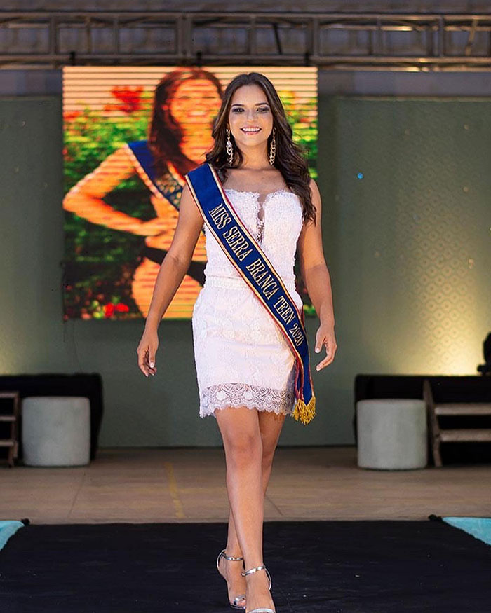 Beauty queen wearing sash and white dress walking on stage at a beauty pageant event with smile.