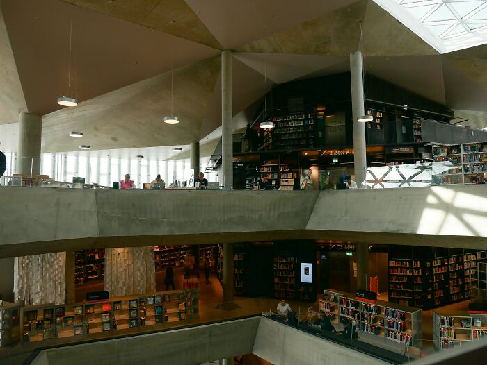 Modern library interior with concrete architecture, shelves filled with books, and visitors reading in a cosy grandiose setting.