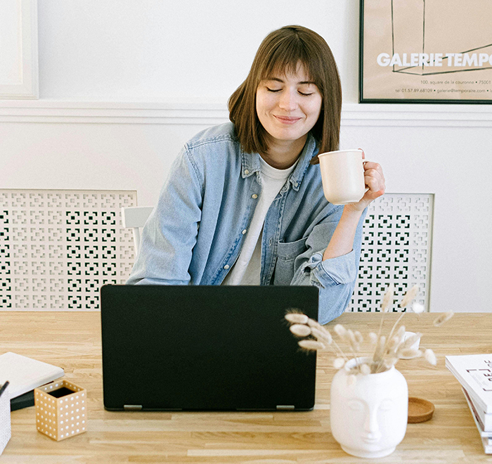 Woman working on laptop at home, smiling while holding a coffee mug, related to job referral bonus story.