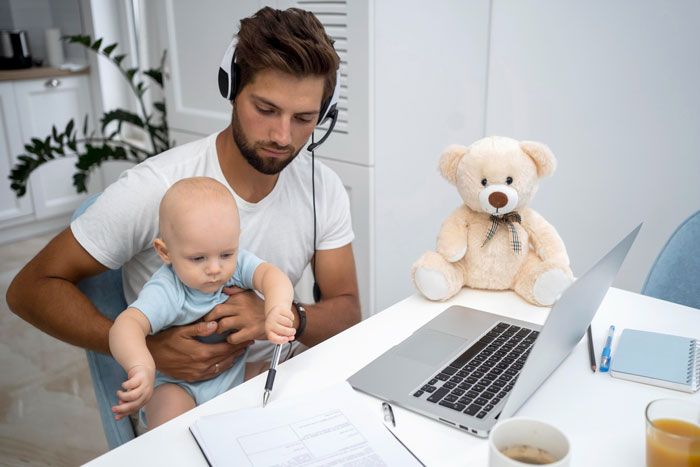 Man babysitting a baby while working on a laptop in a home office showing challenges of real work and babysitting.