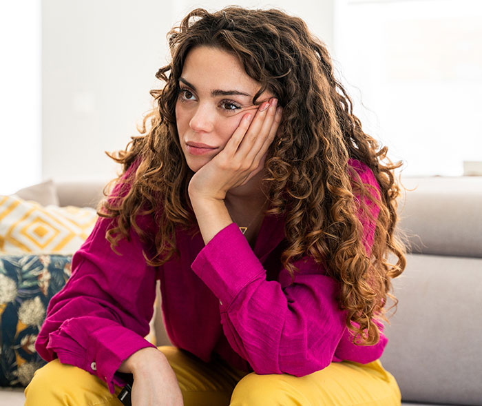 Young woman with curly hair wearing a magenta shirt and yellow pants sitting thoughtfully on a couch indoors.