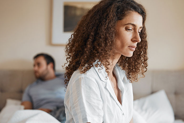 Woman with long curly hair looking upset while boyfriend sits on bed in background refusing to cut her hair for party.