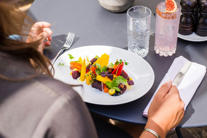 Person sitting at a table with a colorful vegan meal, holding utensils and a glass of water nearby. Person sitting at a table with a colorful vegan meal, holding utensils and a glass of water nearby.
