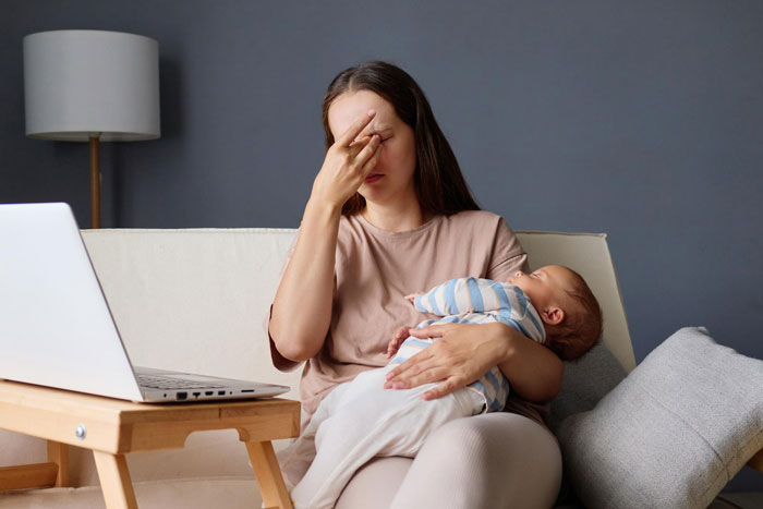 Woman sitting on a couch holding a baby, looking stressed, with a laptop nearby, symbolizing emotions around refusing let rehome cat.